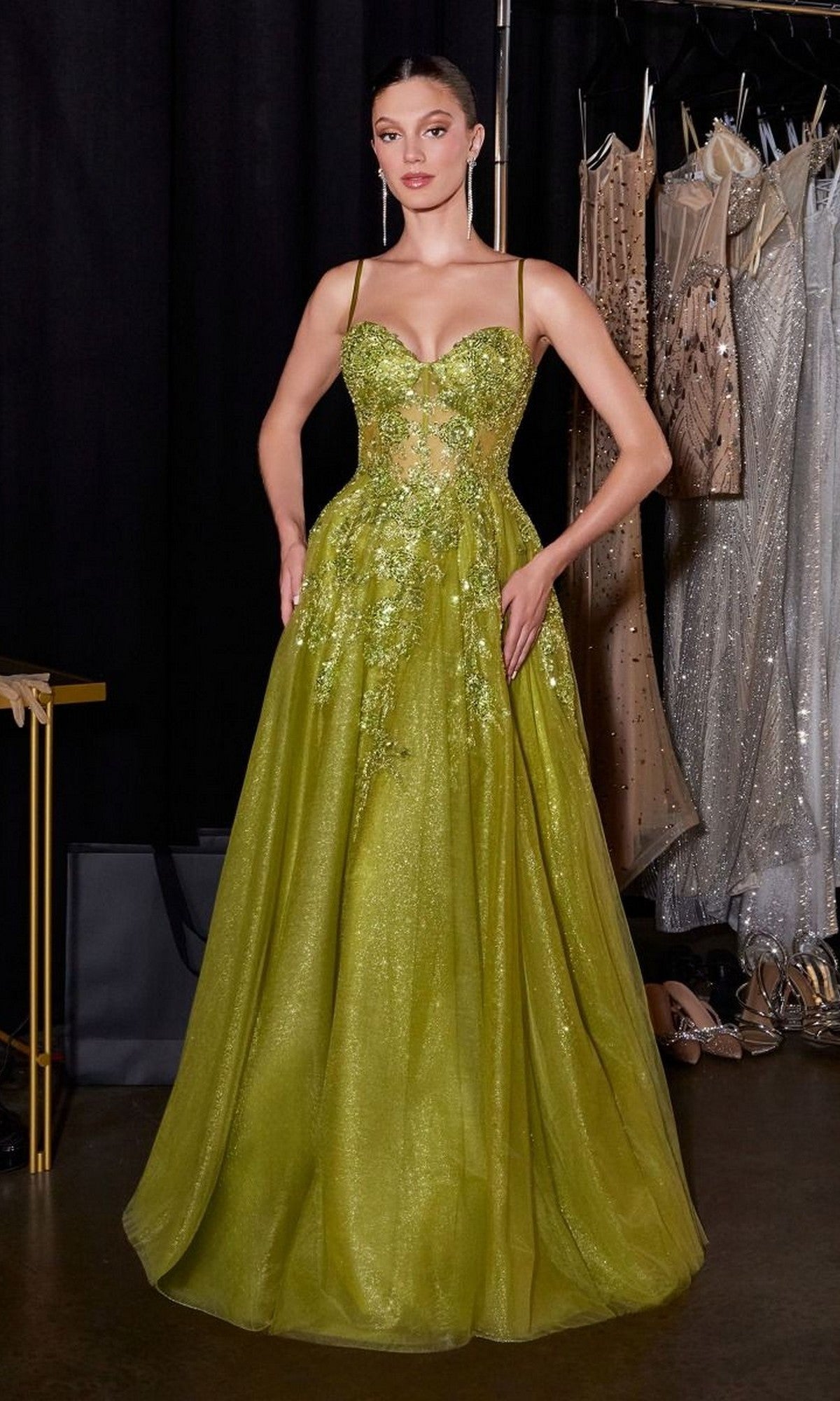 A woman poses backstage in the Ladivine C177, a lime green, floor-length prom dress with thin straps, an embellished bodice, and sequin details, standing before a rack of dresses.